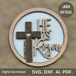 Puede incluir: Un letrero redondo de madera con un fondo azul claro y una cruz de madera marrón con un corazón en el centro. Las palabras "He is Risen" están escritas en una fuente cursiva alrededor de la cruz. Las palabras "Matthew 28:6" están escritas en la cruz. El letrero es perfecto para la decoración de Pascua.