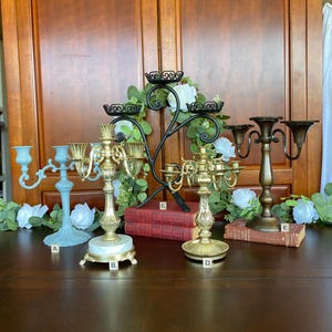 May include: Five metal candelabras labeled A through E. A is light blue, B is gold with a white marble base, C is a black scrollwork design, D is gold, and E is a dark bronze finish. Each piece features multiple arms for holding candles.