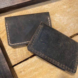 May include: Two dark brown leather cardholders with white stitching are displayed on a light brown wooden surface. The cardholders have a distressed appearance, suggesting a vintage or rustic style. The stitching is visible around the edges.