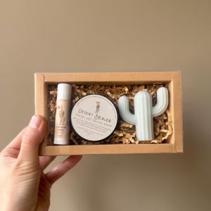 May include: A gift set in a brown box containing a lip balm, a round container labeled "Desert Healer Travel Size Tallow Balm", and a light blue cactus-shaped item. The lip balm has a desert-themed graphic. The box is filled with shredded paper.