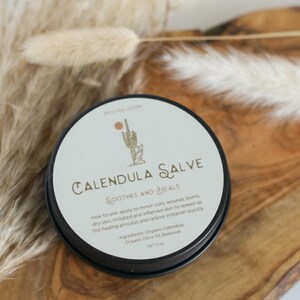 May include: A round black tin of Calendula Salve with a white label featuring a cactus illustration, the text "Calendula Salve Soothes and Heals," usage instructions, and a list of ingredients including organic calendula, olive oil, and beeswax, net 2 oz.