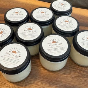 Puede incluir: Nueve frascos pequeños de vidrio transparente llenos de una crema blanca, cada uno con una tapa de metal negro. Una etiqueta blanca circular en cada tapa presenta un gráfico de sol, el texto TALLOW SUN y NATURAL SUN PROTECTION.
