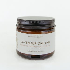 Può includere: Un piccolo barattolo in vetro ambrato con un coperchio a vite in metallo nero. L'etichetta bianca riporta il testo Ancestral Solskin, Lavender Dreams Magnesium Infused Tallow Balm e Handmade in Arizona, con una piccola illustrazione grafica a sinistra.