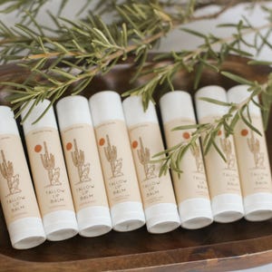 May include: A wooden bowl contains multiple lip balm tubes. Each tube features a beige label with a cactus and sun design, and the words "Tallow Lip Balm." Rosemary sprigs are placed around the tubes.