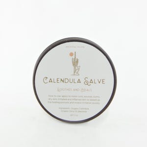 May include: A round container of Calendula Salve with a black lid and a white label. The label features a cactus and coyote illustration, the text Ancestral Solskin, Calendula Salve, Soothes and Heals, usage instructions, ingredients, and Net 2 oz.