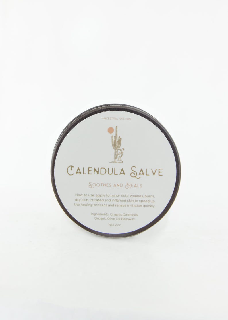 May include: A round container of Calendula Salve with a black lid and a white label. The label features a cactus and coyote illustration, the text Ancestral Solskin, Calendula Salve, Soothes and Heals, usage instructions, ingredients, and Net 2 oz.
