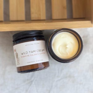 Può includere: Due vasetti di vetro di Wild Yam Cream. Un vasetto è chiuso con un coperchio nero e ha un'etichetta con testo. L'altro vasetto è aperto, rivelando una sostanza cremosa bianco sporco. I vasetti sono su una superficie bianca.