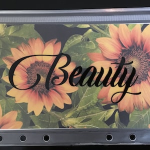 May include: Clear plastic zippered pouch with a floral design featuring sunflowers and green leaves. The word "Beauty" is written in black script across the front. The pouch has a row of reinforced holes along the bottom edge.