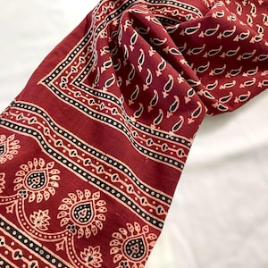 May include: A red cotton scarf with a paisley print and black and white border. The scarf is folded and draped on a white surface.