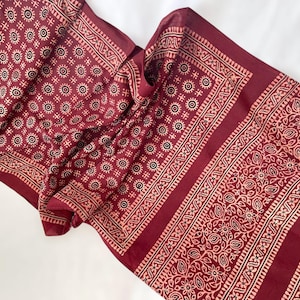 May include: A maroon scarf with a repeating pattern of circular and floral designs in a lighter shade. The scarf has a border with intricate patterns. The fabric is draped, showing the texture and design details. The scarf is likely made of a soft material.