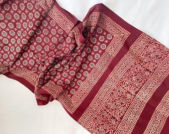 May include: A maroon scarf featuring a repeating pattern of circular and floral designs in a lighter shade. The scarf has a detailed border. The fabric is draped, highlighting the texture and design. The scarf is likely made of a soft material.