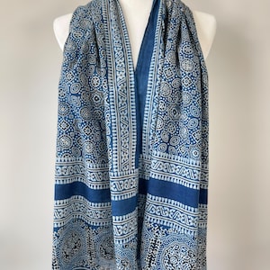 May include: A blue and white patterned scarf draped over a mannequin. The scarf features a repeating geometric design with a border of white and blue patterns. The scarf is made of a lightweight fabric.