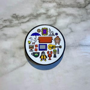 May include: A white circular pin with a black border featuring various icons from the popular TV show "Friends". The icons include a purple door, a couch, a guitar, a penguin, a turkey, a cup of coffee, a lobster, and a duck.