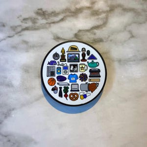 Può includere: Un pop-socket circolare bianco con un bordo nero, con una collezione di piccole illustrazioni colorate. Le immagini includono un pallone da basket, un premio cinematografico, una teiera, un pretzel e una maglietta blu.