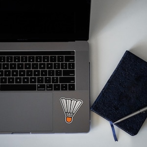 Może przedstawiać: Srebrny laptop z naklejką białego i pomarańczowego lotka do badmintona na touchpadzie. Po prawej stronie laptopa znajduje się ciemnoniebieski notes z długopisem.