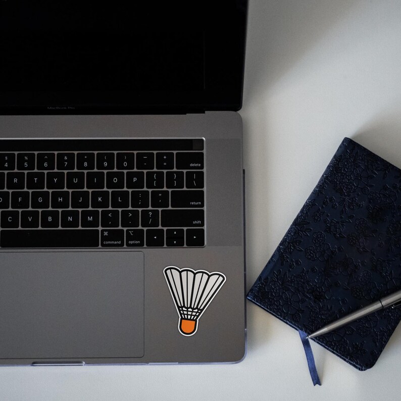 Może przedstawiać: Srebrny laptop z naklejką białego i pomarańczowego lotka do badmintona na touchpadzie. Po prawej stronie laptopa znajduje się ciemnoniebieski notes z długopisem.