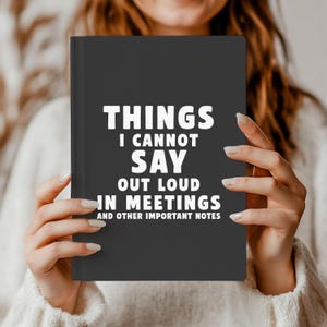 May include: A dark grey notebook featuring the white text "THINGS I CANNOT SAY OUT LOUD IN MEETINGS AND OTHER IMPORTANT NOTES". The notebook is held by a person.
