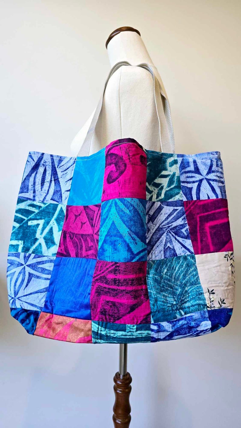 May include: A large patchwork tote bag with a variety of blue, pink, and green patterns. The bag has a white canvas strap.