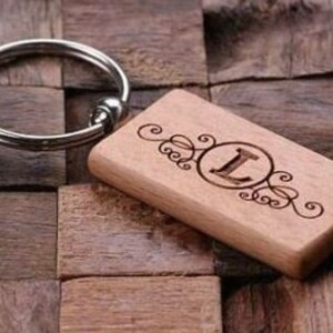 May include: A wooden keychain with a silver key ring and a rectangular wooden tag with the letter "L" engraved in a circle surrounded by a decorative design.