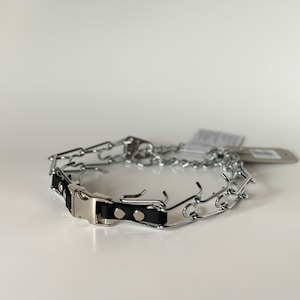 May include: A silver prong collar for dogs with a black leather strap and a silver buckle. The collar is lying on a white surface.