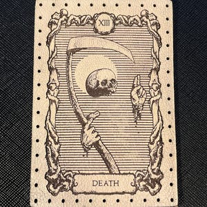 May include: A tarot card featuring a detailed illustration of the Death card. The card depicts a skeletal figure holding a scythe, a skull, and a hand. The word "DEATH" is at the bottom, with the Roman numeral "XIII" at the top.