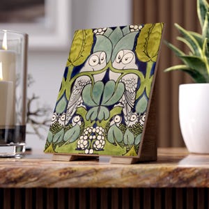 May include: A ceramic tile with a green, blue and white design featuring two owls and a floral pattern. The tile is mounted on a wooden stand.