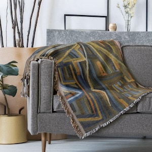 May include: A woven throw blanket with a geometric design in shades of olive green, blue, orange, and yellow is draped over a gray sofa. The blanket has a fringed edge. The sofa has wooden legs and is placed against a neutral background.
