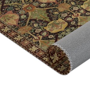 Puede incluir: Una alfombra enrollada con un dise&ntilde;o estampado en marr&oacute;n oscuro, verde y dorado. La alfombra tiene un borde con flecos y est&aacute; enrollada sobre un respaldo gris.