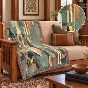 May include: A woven throw blanket featuring a geometric patchwork pattern in shades of teal, olive green, mustard yellow, and cream. The blanket has a textured surface with visible thread details and a fringed edge, draped over a light-colored sofa.