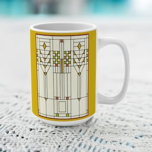 May include: A white ceramic mug featuring a mustard yellow background and a central rectangular panel with a geometric stained glass pattern in cream, green, and orange tones, composed of vertical lines, diamonds, and small square grid accents.