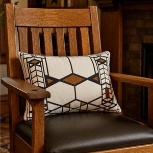 May include: A rectangular throw pillow featuring a white background with a black geometric grid pattern, accented by brown and red diamond and rectangular shapes, resting on a dark brown leather seat of a wooden chair with vertical slats.