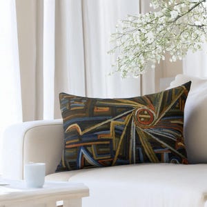 May include: Rectangular decorative pillow with an abstract geometric design in shades of brown, blue, orange, and yellow. The pillow is placed on a white sofa, with a white table and a vase of white flowers in the background.