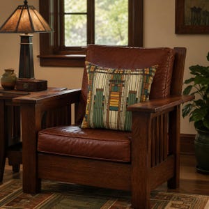 May include: A square throw pillow featuring a geometric pattern of vertical lines and rectangular blocks in shades of cream, forest green, mustard yellow, and dark brown, resting on a dark brown leather armchair with a wooden frame.