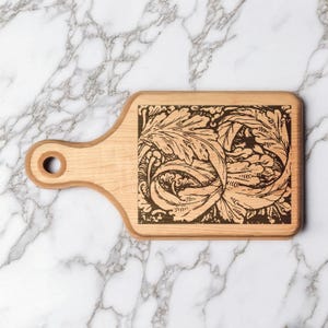 Puede incluir: Una tabla de cortar de madera clara con un asa y un agujero circular. La superficie rectangular presenta un diseño floral oscuro grabado. La tabla está sobre un fondo de mármol.