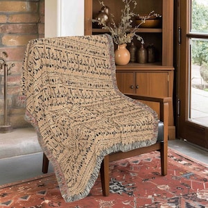 May include: A woven throw blanket featuring a repeating pattern of dark brown and black geometric shapes on a tan background. The blanket has a textured surface with a fringed edge along the bottom and sides, draped over a wooden chair.