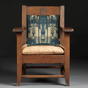 May include: A square throw cushion featuring a geometric pattern in shades of blue, cream, and brown, resting on a dark brown wooden armchair with a light tan seat cushion. The cushion design consists of vertical lines and angular shapes.
