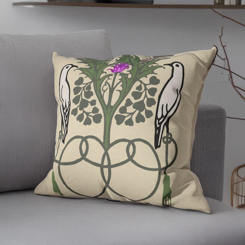 Pu&ograve; includere: Un cuscino quadrato con sfondo beige caratterizzato da un design grafico simmetrico. Due uccelli bianchi sono rivolti verso una pianta verde centrale con un fiore viola. Il disegno include linee intrecciate verde scuro e nappe.