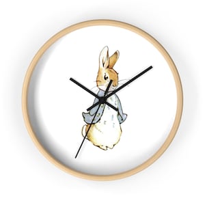 May include: A round wall clock with a light wood frame. The clock face features a watercolor illustration of a rabbit wearing a blue jacket. Black clock hands indicate the time. The background is white.