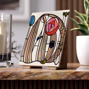 May include: A decorative ceramic tile with a floral design in shades of pink, red, blue, and brown, set on a wooden stand. The artwork features a stylized rose and abstract shapes, reminiscent of stained glass. The tile is displayed on a marble-like surface.