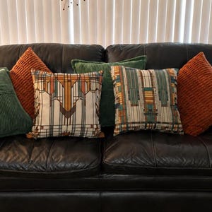 May include: A collection of square throw pillows arranged on a dark leather sofa. The set includes two solid orange corduroy pillows, two solid green corduroy pillows, and two pillows featuring a geometric pattern in shades of orange, green, and tan.