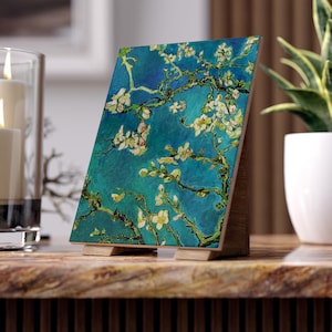 Può includere: Un supporto in legno con una piastrella quadrata che presenta un dipinto di un mandorlo in fiore con fiori bianchi su uno sfondo blu. Il dipinto è in stile Vincent van Gogh.