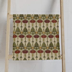 May include: A decorative towel with a repeating floral pattern in red, green, and yellow on a cream background. The towel is draped over a wooden ladder, showcasing the Art Nouveau-inspired design. The pattern features stylized flowers and leaves.