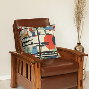 K&ouml;nnte beinhalten: Ein brauner Ledersessel mit einem dekorativen Kissen mit einem farbenfrohen geometrischen Design. Das Kissen hat rote, blaue, schwarze und wei&szlig;e Formen. Der Text "Frank Lloyd Wright" befindet sich oben im Bild.