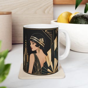 May include: A white ceramic mug featuring a black and gold graphic illustration of a person wearing a cloche hat and a dress, set against a stylized skyscraper background with geometric fan patterns in a distinct art deco inspired aesthetic.