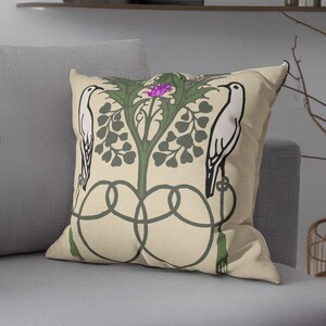 Pu&ograve; includere: Un cuscino quadrato con sfondo beige caratterizzato da un design grafico simmetrico. Due uccelli bianchi sono rivolti verso una pianta verde centrale con un fiore viola. Il disegno include linee intrecciate verde scuro e nappe.
