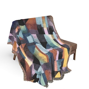 以下が含まれることがあります： 青、オレンジ、黄色、紫の色合いの幾何学模様が施された、カラフルな織りのスローブランケット。 ブランケットにはフリンジエッジがあり、茶色のベンチに掛けられています。