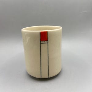 May include: A cylindrical ceramic cup with an off-white, smooth finish. The exterior features a single vertical line design in black ink, topped with a small, solid rectangular block of vibrant orange color near the rim of the vessel.