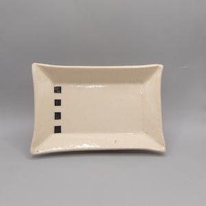 May include: A rectangular ceramic dish with a cream-colored glaze. Four small black squares are arranged vertically on the left side. The dish has slightly curved edges and a minimalist design, suitable for serving food or as a decorative piece.