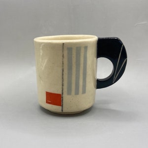 May include: Cream-colored ceramic mug with a dark blue handle. The mug features vertical gray stripes and a red square with a horizontal line. The handle has a unique, curved design.
