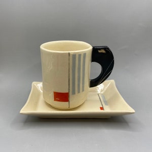 May include: Cream-colored ceramic mug and saucer set. The mug has a black handle and is decorated with vertical gray stripes and a red square. The square saucer has matching design elements. A modern, minimalist design.